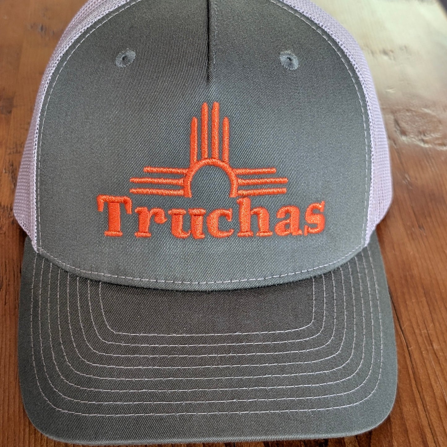An army green trucker hat with a curved brim, featuring an orange embroidered Truchas logo and a Zia sun symbol on the front. The back of the cap has light gray mesh panels, and the hat is photographed on a wooden surface.