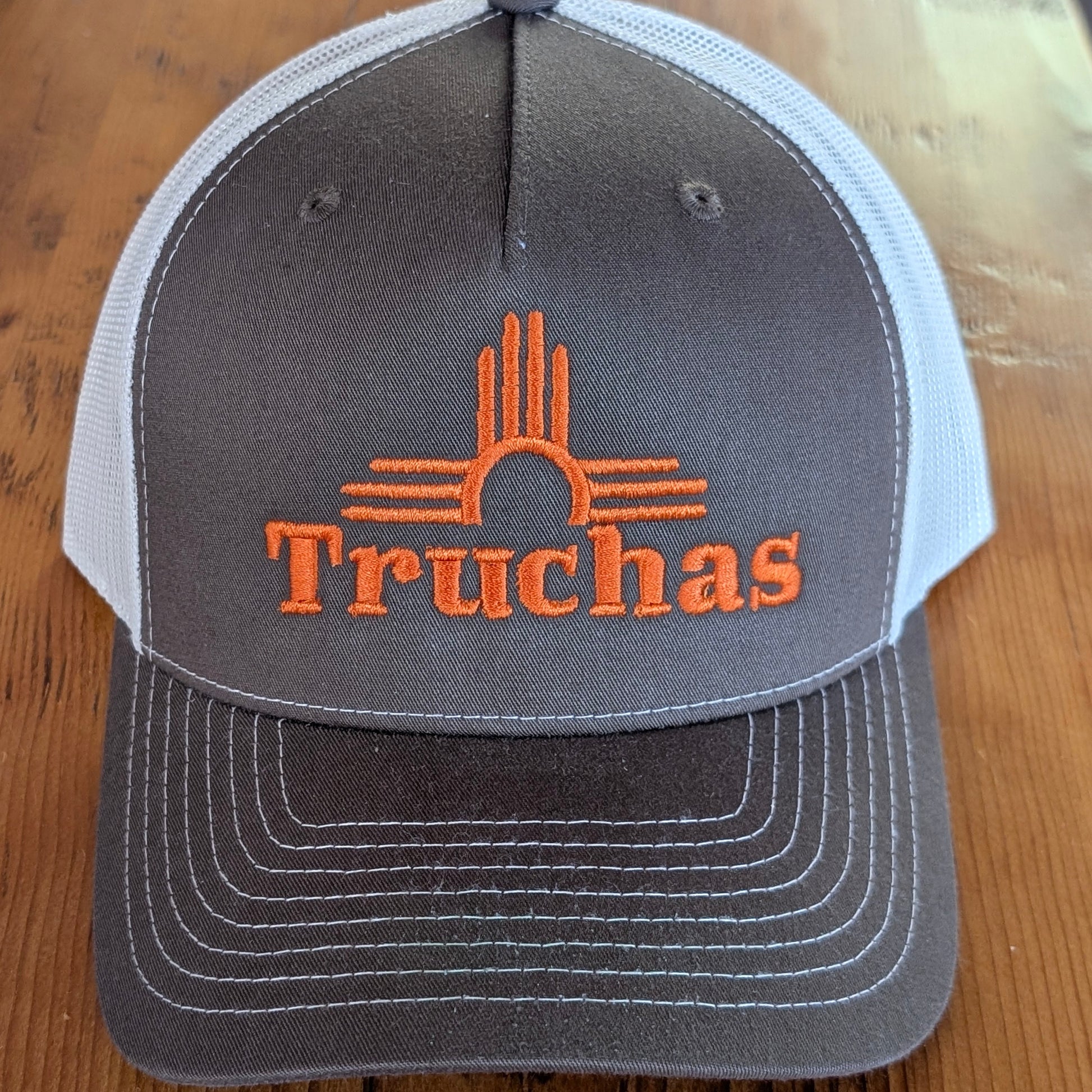 A chocolate brown and white trucker hat with an orange embroidered Zia and the word 'Truchas' on the front, resting on a wooden surface.